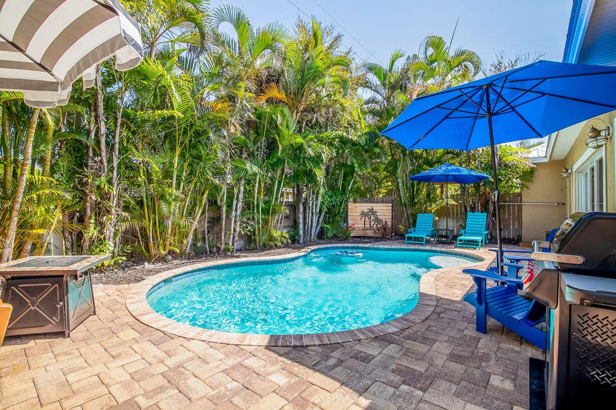 Private freeform heated pool and tropical paver patio at Coastal Cottage, a 3-bedroom vacation home in Holmes Beach on Anna Maria Island. Lush palm landscaping creates a secluded backyard oasis with lounge seating, shaded umbrellas, and a gas grill — perfect for Gulf Coast days just two block from the beach.