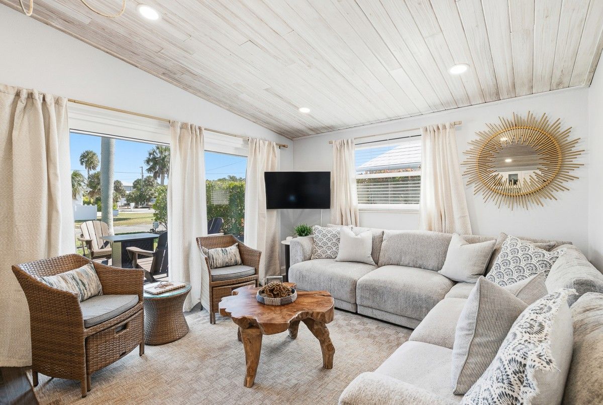 Bright living room with a gray sectional, wicker chairs, and a natural wood coffee table under a light, airy ceiling and sunburst mirror.